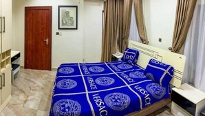 A bedroom with a bed covered in a blue and white patterned bedspread, a wooden wardrobe, a small bedside table, and a framed picture on the wall. - House - Sleeps 6 & private parking (Ikeja GRA)