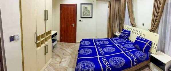 A bedroom with a bed covered in a blue and white patterned bedspread, a wooden wardrobe, a small bedside table, and a framed picture on the wall. - House - Sleeps 6 & private parking (Ikeja GRA)