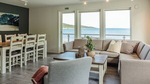 A modern living room with a dining area, large windows with a view of the sea, and a painting on the wall.