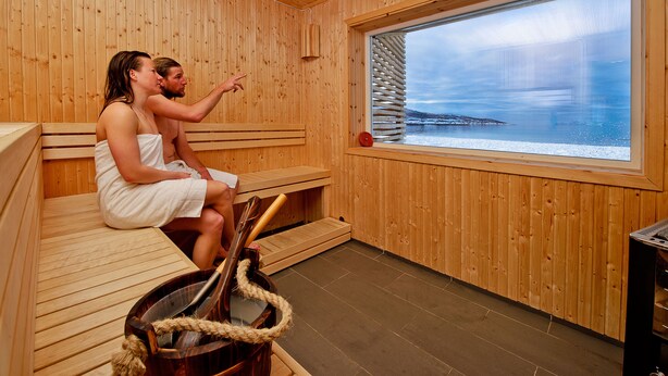A sauna with wooden walls and benches, a large window with a view of a snowy landscape, and a sauna brush and rope.