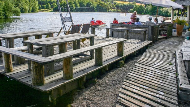 Een houten steiger met picknicktafels en een boot op de achtergrond.