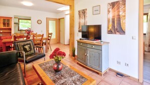 Een ruime woonruimte met een houten plafond, een eettafel met stoelen, een bank, een televisie op een kast en een bloemenvaas.