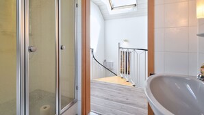 Een badkamer met een douchecabine van glas, een houten deur, een wit wastafelmeubel en een dakraam boven de douche.