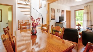Een eethoek met een houten tafel en stoelen, een vaas met bloemen, een televisie op een kast, en een trap naar een andere kamer.