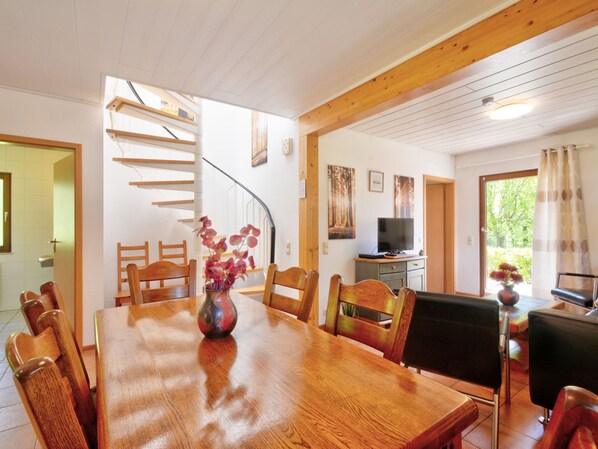 A dining area with a wooden table and chairs, a vase with flowers, a television on a cabinet, and a staircase leading to another room.