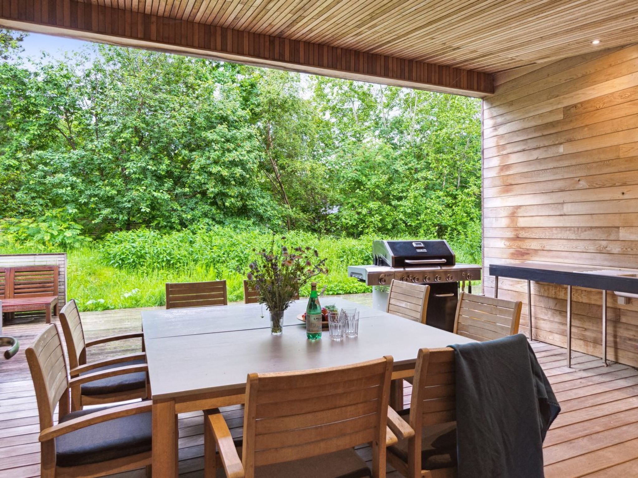 Eine Terrasse im Freien mit Holztisch und -stühlen, einem Grill und Blick auf einen üppigen grünen Wald.