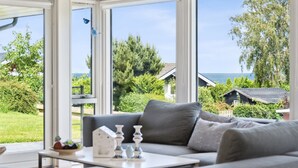 Ein Wintergarten mit grauem Sofa, einem kleinen Tisch und Dekoration. Große Fenster bieten Blick auf Meer und Grünflächen.
