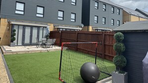 A residential backyard with a fenced play area, a patio with outdoor furniture, and a modern two-story house.