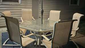En udendørs terrasse med et glasbord og fletstole, under en stor parasol med integreret lys.