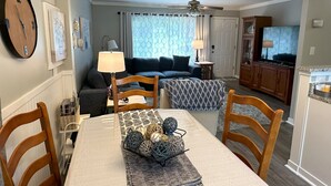 Spiseområde med et bord dækket til fire personer, en sofa, en loftventilator og et fjernsyn i baggrunden.