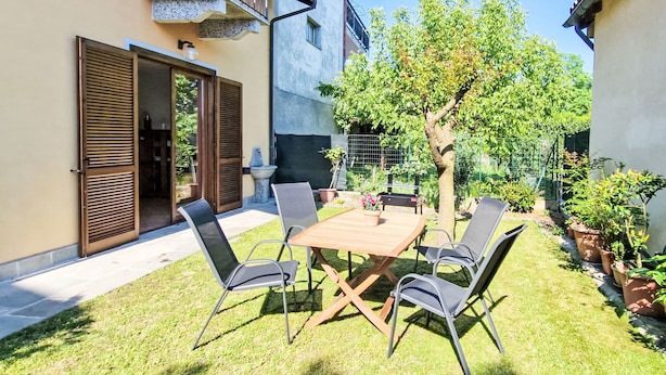 A patio with a wooden table and four chairs, surrounded by a well-maintained lawn and potted plants. - Sky & Lake by Interhome (Arizzano)