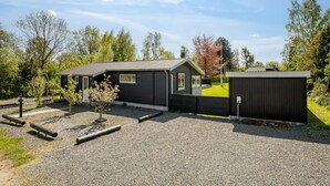 En modern byggnad i två våningar med en mörk fasad, en liten veranda och en inhägnad trädgård med grusgångar.