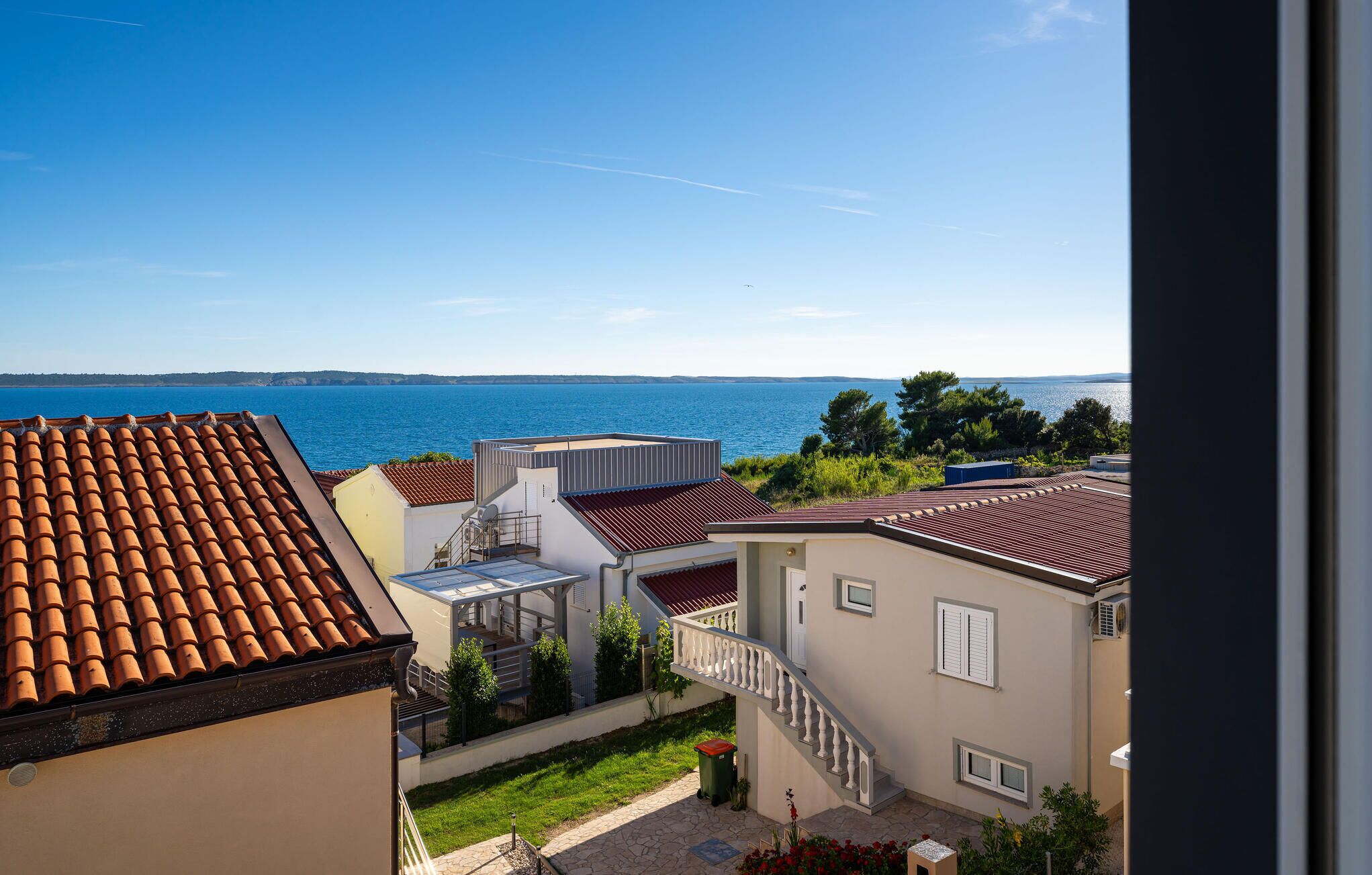 Un quartier résidentiel offrant une vue sur la mer, caractérisé par des toits recouverts de tuiles en terre cuite, un bâtiment blanc doté d’un balcon et une poubelle verte.