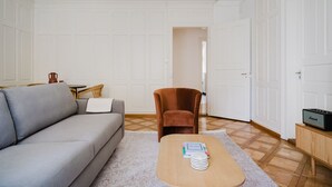 Olohuoneessa on harmaa sohva, ruskea nojatuoli, pieni puinen pöytä, jonka päällä on kirjoja ja pino lautasia, sekä Marshall-kaiutin puisen kaapin päällä.