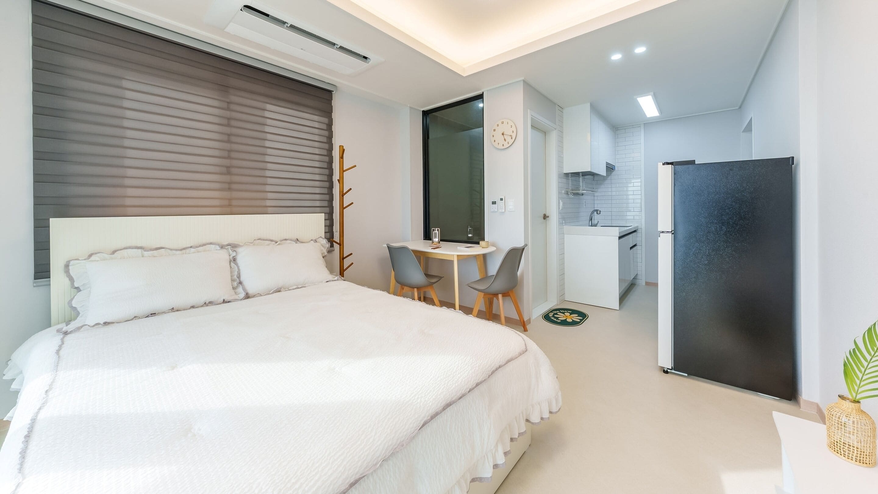 A modern hotel room with a large bed, a desk with two chairs, a small kitchenette, and a refrigerator.