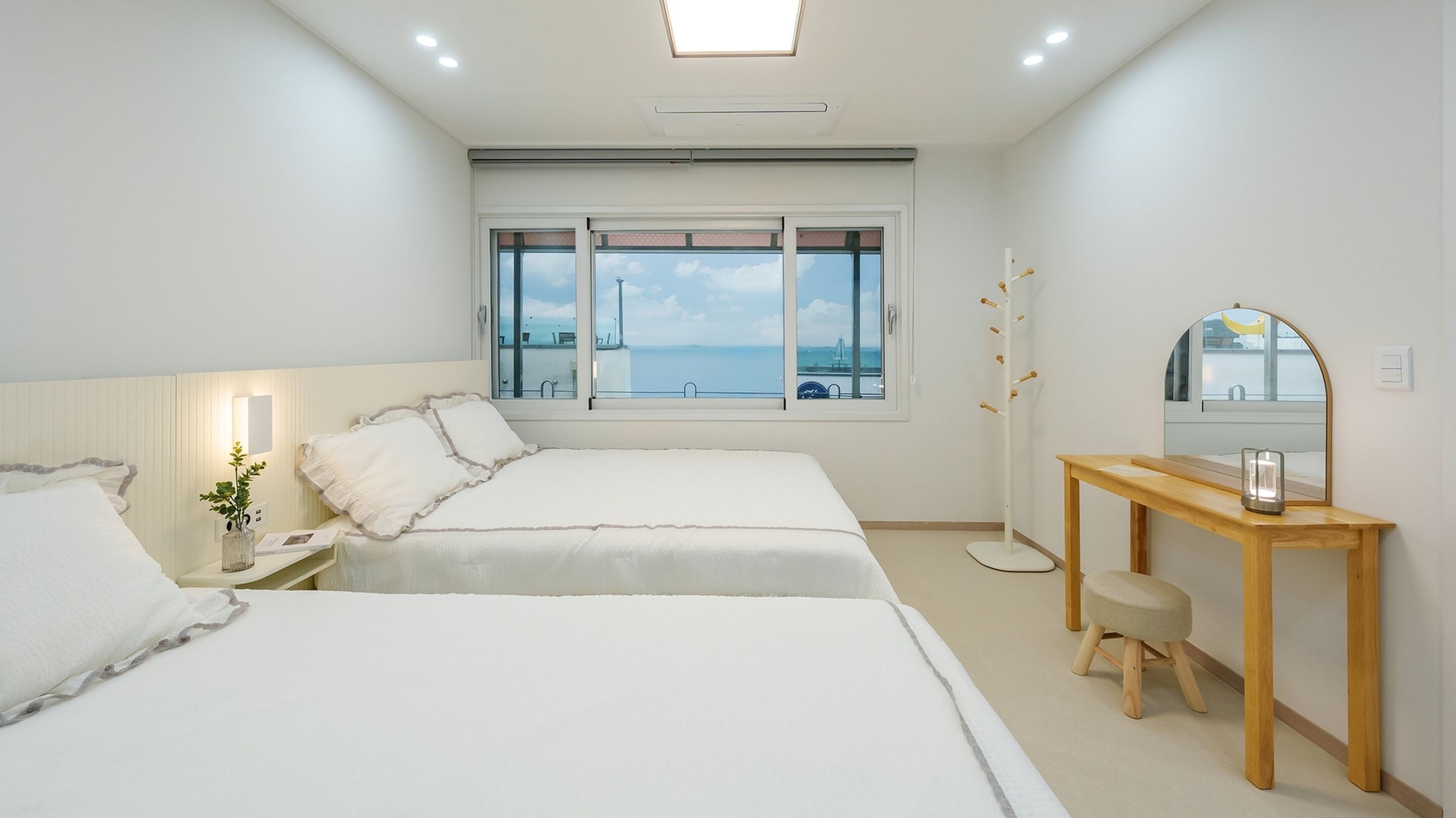 A hotel room with two beds, a wooden nightstand, a mirror, and a view of the sea through large windows.