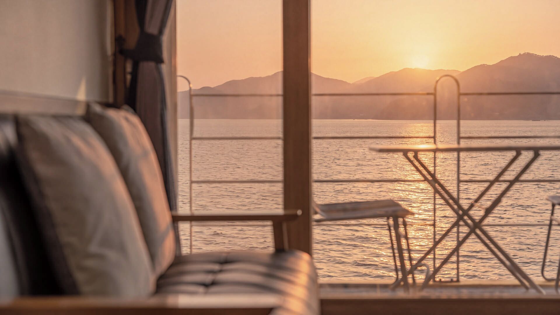 A view from a room with a window overlooking a body of water and mountains during sunset.
