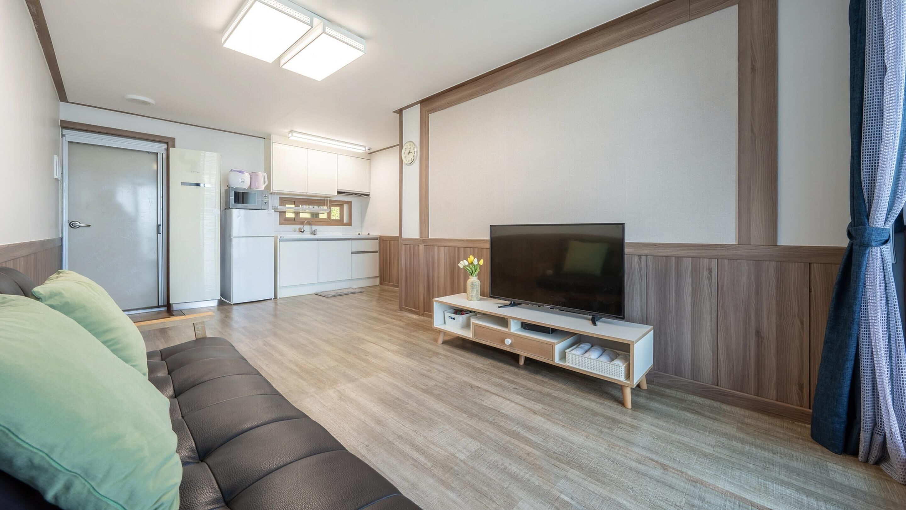 A compact living space with a sofa, a TV on a wooden stand, a kitchenette, and a door leading to a bathroom.