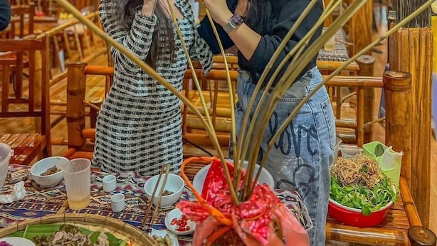 A restaurant interior with wooden furniture, a central table with food, and a decorative plant arrangement. - Mami Home - Homestay Lac Village (Mai Ha)