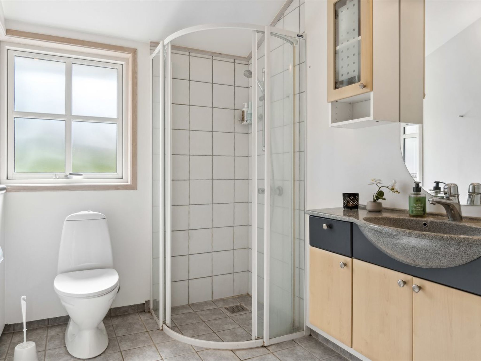 A bathroom with a toilet, a shower enclosure, a sink with a mirror, and a window.