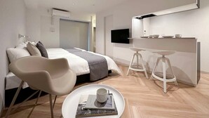 A modern hotel room with a bed, a desk with two stools, a TV, and a small table with a cup and book. - Breakfast & Comfort in Hiroshima's Heart (Naka Ward, Hiroshima)