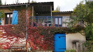 A house with a blue door, a balcony, and a garden area with a bench and hose. - La Petite Maison D'omélie (TOURNEMIRE)