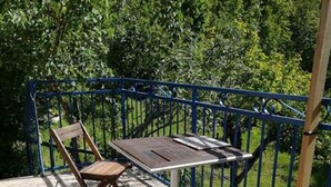 A wooden table and chairs on a concrete patio with a blue metal railing, surrounded by greenery. - La Petite Maison D'omélie (TOURNEMIRE)