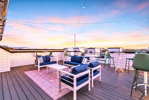 Um deck externo com móveis azuis e brancos, luzes de corda e vista do horizonte da cidade ao pôr do sol.