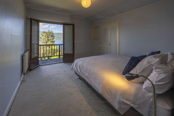 A bedroom with a bed, a nightstand, a lamp, a door, and a balcony with a view of trees and hills.