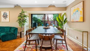 A dining area with a large wooden table, six chairs, and a green sofa. There are potted plants and a painting on the walls. - 6BR Villa with Pool, Jacuzzi & Game Room, Walk to Rodeo Drive (Los Angeles)