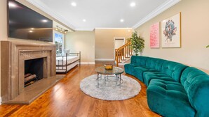 A living room with a fireplace, a large flat-screen TV, a teal sofa, and a staircase leading to another level. - 6BR Villa with Pool, Jacuzzi & Game Room, Walk to Rodeo Drive (Los Angeles)