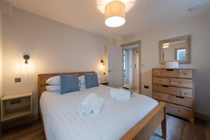 Een slaapkamer met een houten bed, witte lakens, twee wandlampen, een houten dressoir en een spiegel.