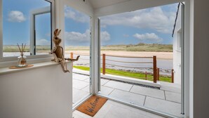 Een kamer met uitzicht op een strand en duinen, een houten konijnenbeeldje op de vensterbank, en een welkomstmat bij de deur.