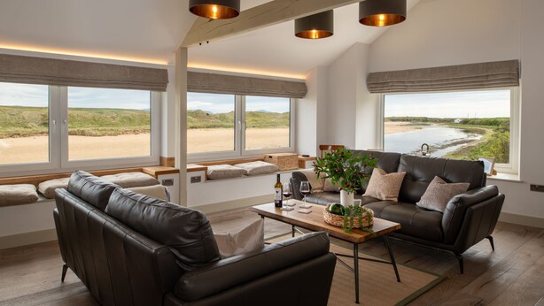 Een ruime woonkamer met een leren bank, een houten salontafel en uitzicht op een rivier en heuvels.