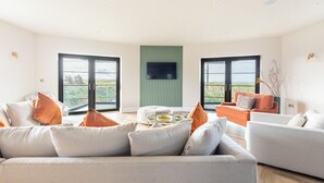 A modern living room with a sofa, armchairs, and a coffee table. There are large windows with a view of the outdoors. - Bluebells: Eco-Friendly 4-Bed Home with Sea Views in Llanfaelog | Managed by Anglesey Holiday Lettin (Llanfaelog)