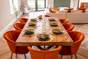 Mesa de comedor preparada para seis personas con sillas anaranjadas, rodeada por una zona de estar moderna con un sofá y un ventanal.