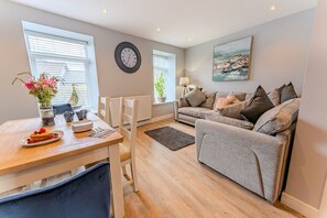 A modern living room with a sofa, a dining table with a vase of flowers, and a clock on the wall. - No. 66: Stylish Pet-Friendly Cottage in Beaumaris (Beaumaris)