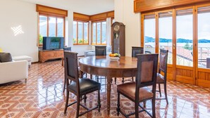 A dining area with a round wooden table, six chairs, a clock, and a television on a cabinet. Large windows offer a view of the mountains.