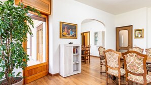 A spacious living room with wooden flooring, a large potted plant, a white entertainment unit, and a dining area with floral-patterned chairs.