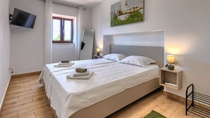 Un lit double bien fait, avec du linge de lit blanc, un téléviseur à écran plat fixé au mur, une fenêtre avec des rideaux, un tableau au-dessus du lit et une table de chevet avec une lampe.