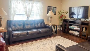 A living room with a dark leather sofa, a TV on a stand, a lamp, a small table, and a decorative cabinet.