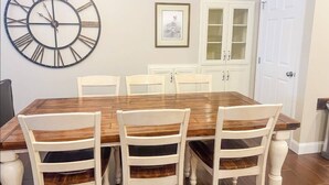 A dining room with a wooden table, four chairs, a large wall clock, a framed picture, and white cabinets.
