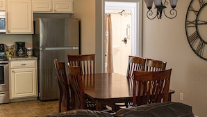 A kitchen with white cabinets, a stainless steel refrigerator, a dining table with wooden chairs, and a wall-mounted clock.