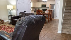 A living room with a leather sofa, a dining table with chairs, a kitchen area with cabinets and appliances, and a staircase.