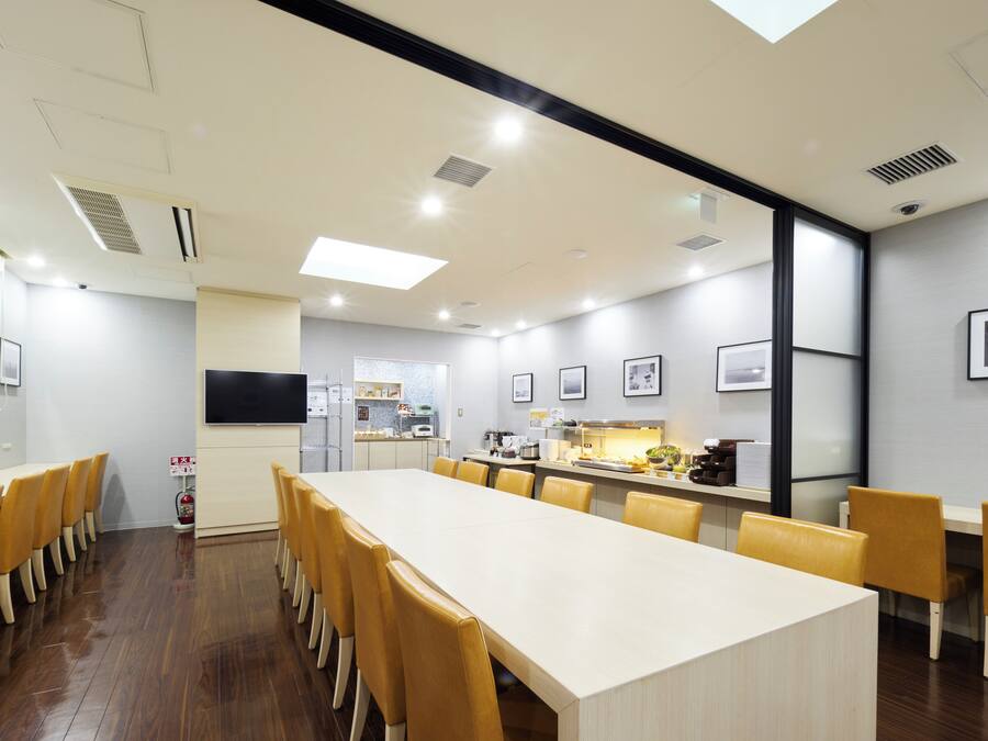 Uma sala de conferência moderna com uma mesa comprida e cadeiras amarelas, uma pequena cozinha ao fundo e um extintor de incêndio na parede.