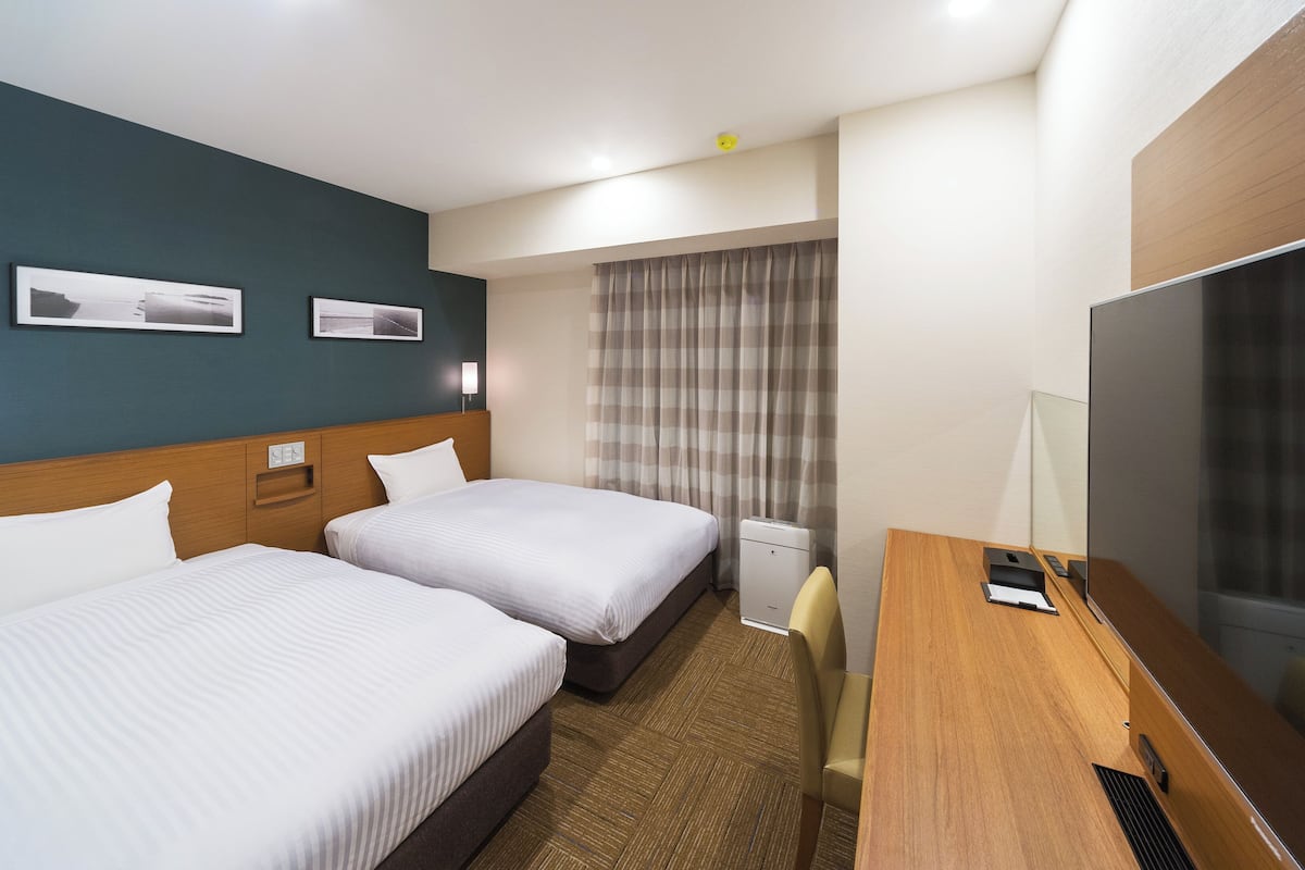 Habitación de hotel con dos camas, un escritorio, un televisor de pantalla plana, una silla, una ventana con cortinas y una lámpara fijada a la pared.