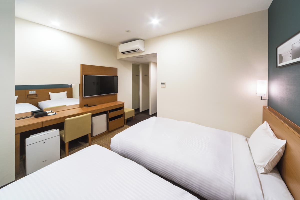 Habitación de hotel con dos camas, un escritorio con televisor, un refrigerador pequeño y aire acondicionado instalado en la pared.