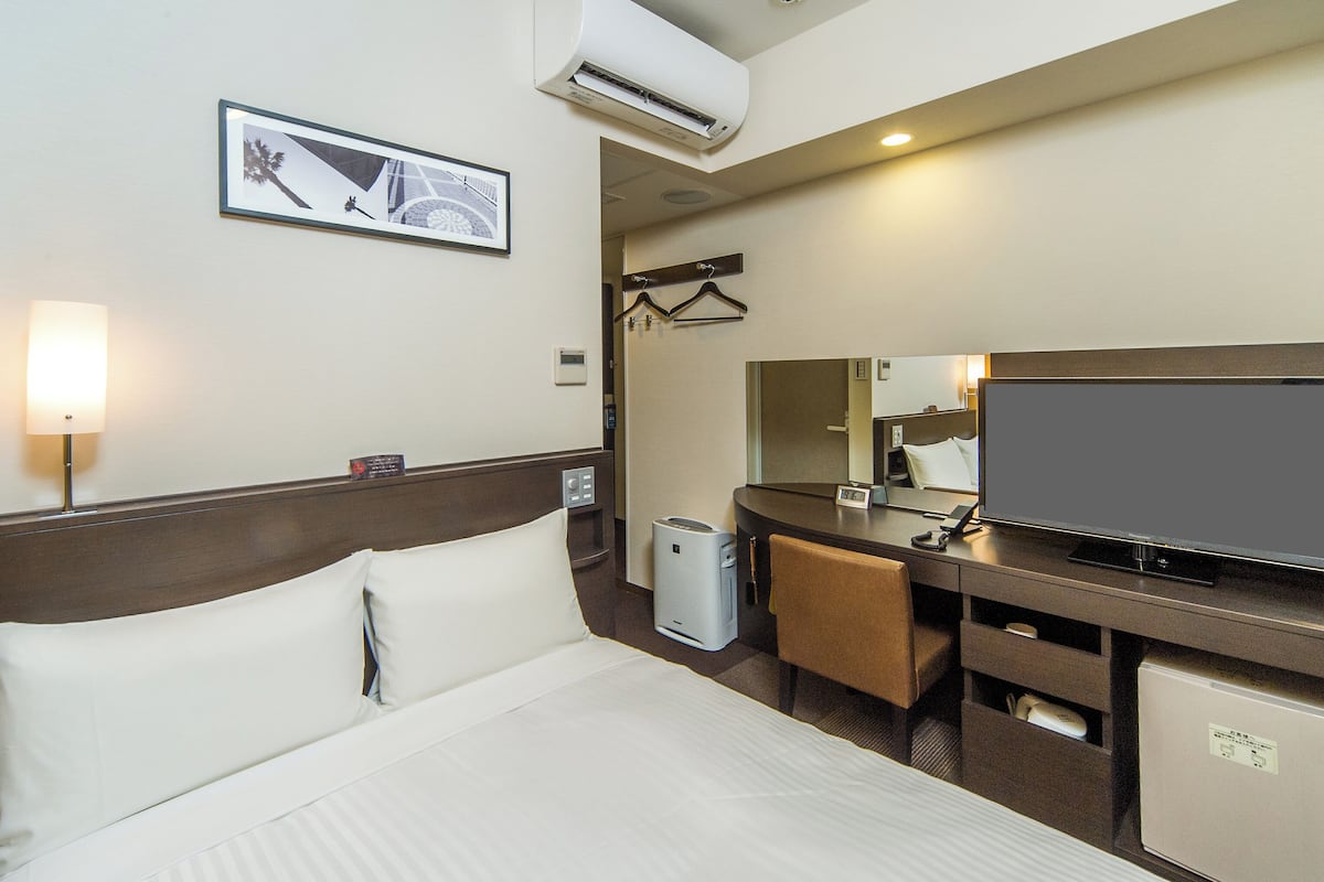Habitación de hotel con cama, escritorio con televisor de pantalla plana, espejo, secador de pelo y un refrigerador pequeño.