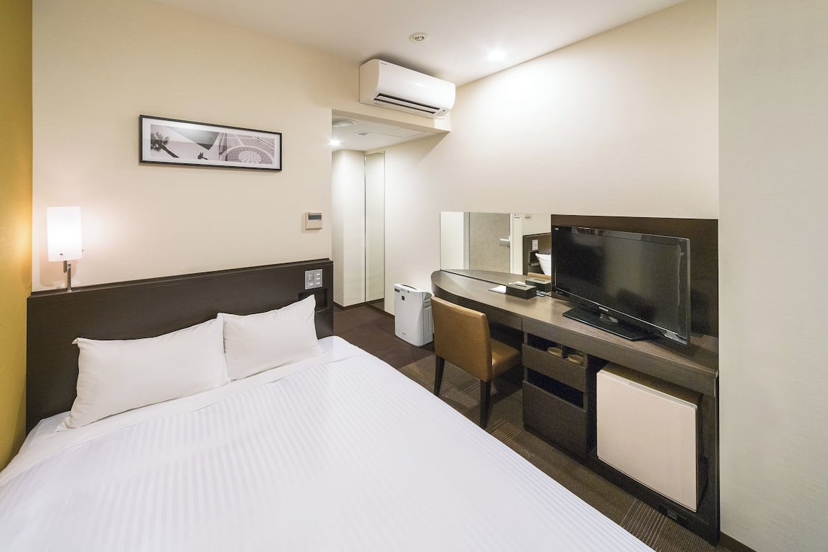 Una habitación de hotel con una cama grande, un escritorio con televisor, una silla y un equipo de aire acondicionado.