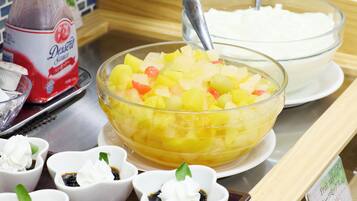 Una mesa de buffet con un bol grande de ensalada de frutas, recipientes pequeños de crema batida y una botella de salsa para postres.